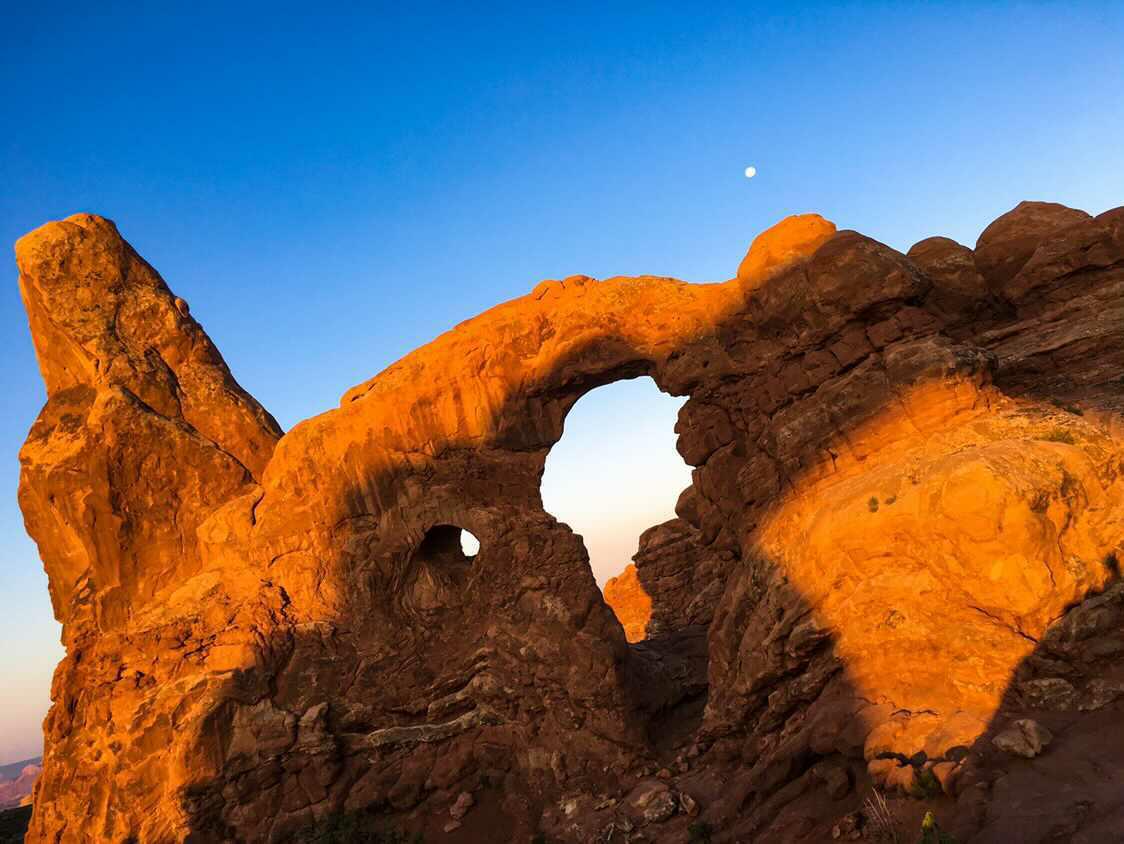 Turret Arch glowing orange at sunrise with moon above
